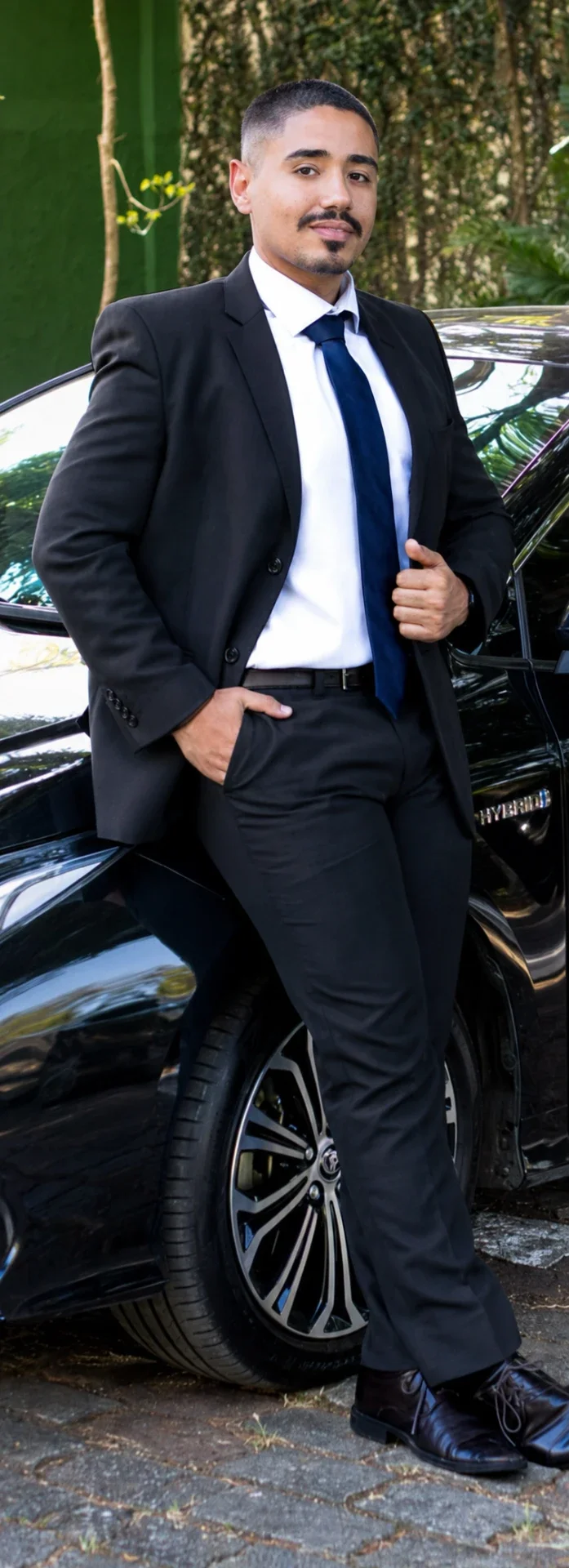 Gustavo Gomes, fundador da Execdriver Academy, de terno preto e gravata ao lado de um sedã executivo preto.
